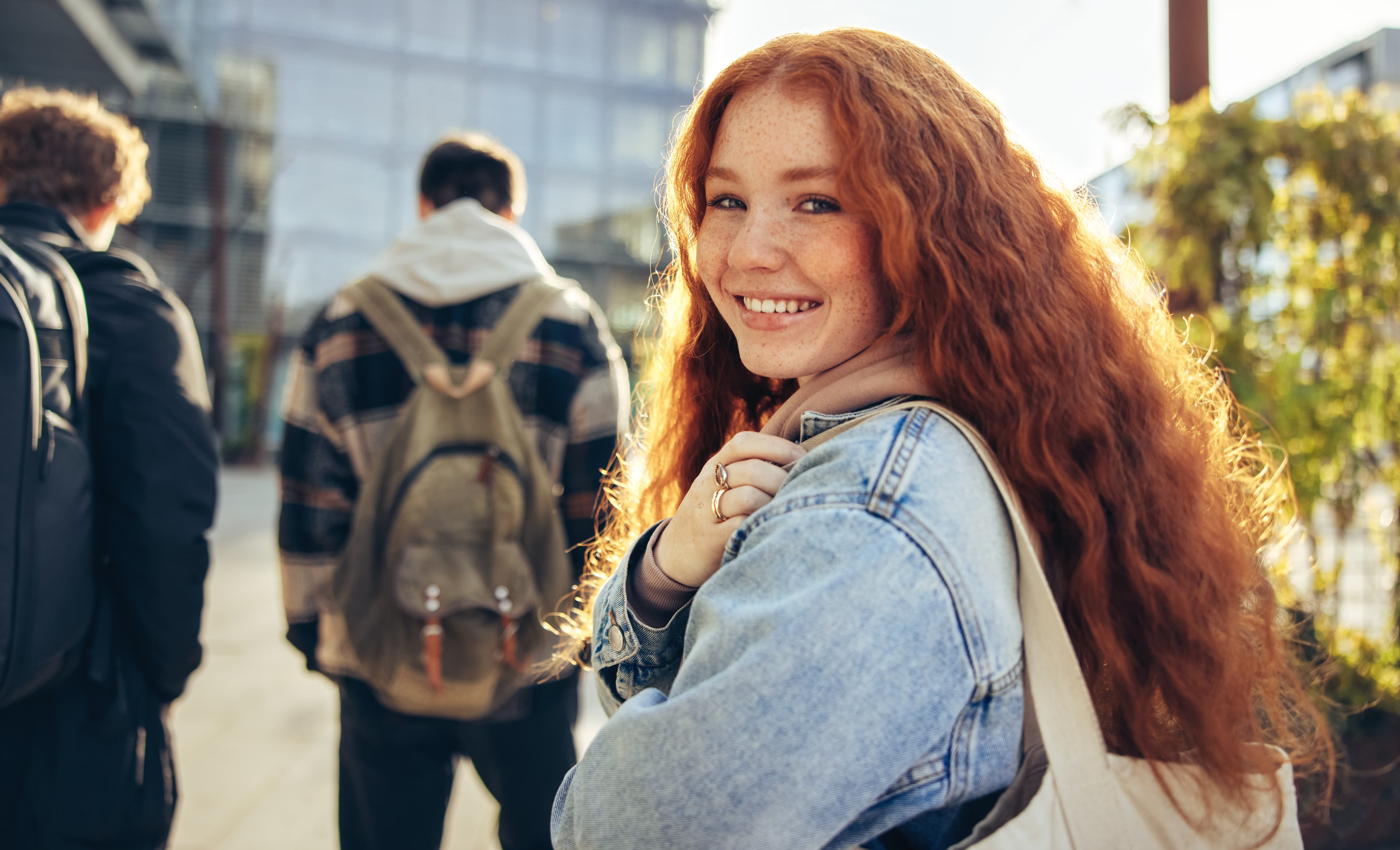A young woman with red hair and a denim jacket is smiling and looking over her shoulder. She is carrying a tote bag.